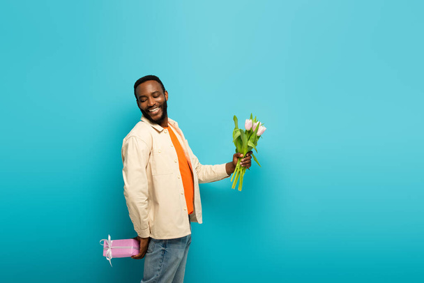 cheerful african american man holding tulips and while hiding present behind back on blue - Foto, Imagem