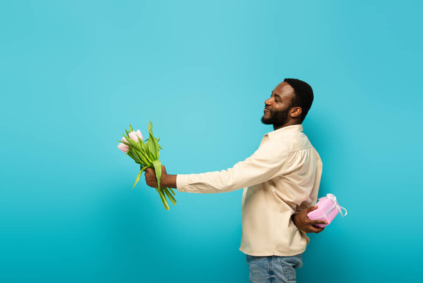 sonriente afroamericano hombre presentando tulipanes mientras oculta caja de regalo detrás de nuevo en azul  - Foto, imagen