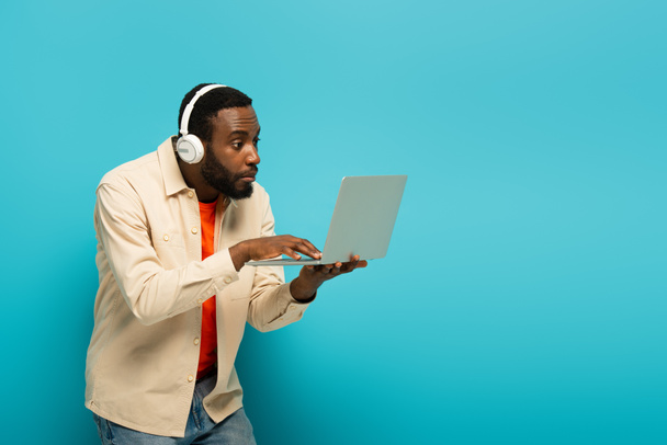 překvapený africký Američan ve sluchátkách pomocí notebooku na modrém pozadí - Fotografie, Obrázek