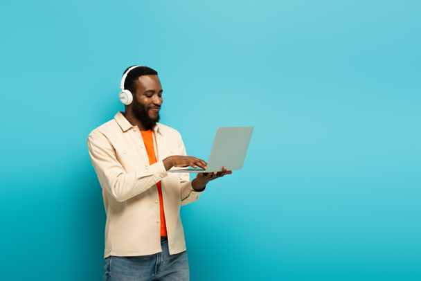 pleased african american man in headphones typing on laptop on blue background - Foto, afbeelding