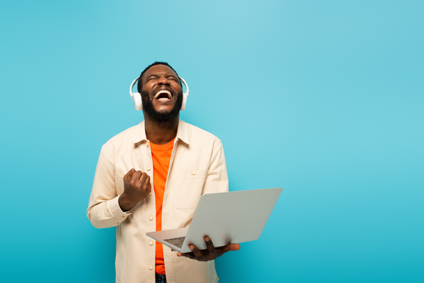 podekscytowany african amerykański człowiek w słuchawki pokazując gest sukcesu podczas stania z laptopem izolowane na niebiesko - Zdjęcie, obraz