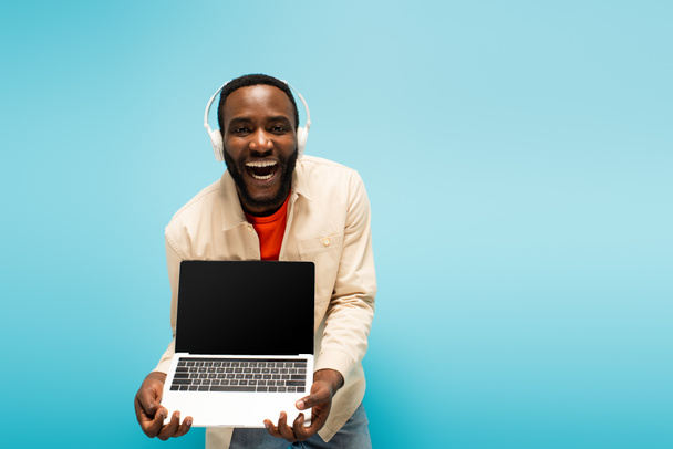 asombrado hombre afroamericano en auriculares que muestran portátil con pantalla en blanco aislado en azul - Foto, imagen
