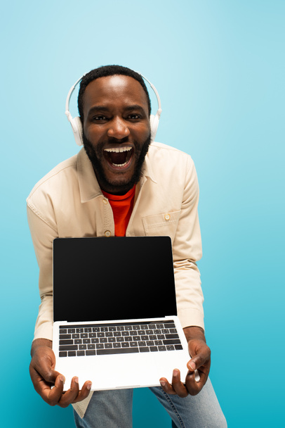 smějící se africký Američan ve sluchátkách ukazující notebook s prázdnou obrazovkou na modré  - Fotografie, Obrázek