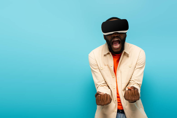 angry african american man in vr headset screaming and showing clenched fists isolated on blue - Zdjęcie, obraz