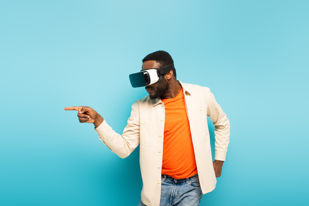 junger afrikanisch-amerikanischer Mann zeigt mit dem Finger zur Seite, während er im vr-Headset auf blauem Hintergrund spielt - Foto, Bild