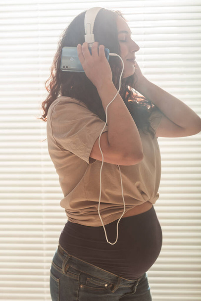 Curly-haired brunette pacified pregnant woman listens to music using smartphone and headphones. Concept of a soothing mood before meeting baby. - Photo, Image