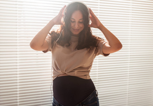 Χαμογελώντας μελαχρινή γαλήνια έγκυος γυναίκα ακούγοντας ευχάριστη μουσική χρησιμοποιώντας ένα smartphone και ακουστικά. Έννοια θεραπεία καταστολή πριν από τη συνάντηση με το μωρό - Φωτογραφία, εικόνα
