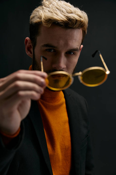módní muž drží brýle v rukou a bunda oranžový svetr portrét zblízka - Fotografie, Obrázek