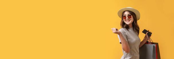 Bella donna asiatica shopaholic sorride mentre trasporta borse della spesa e smartphone su sfondo giallo. Summer sale, Concetto banner di vendita a metà anno. - Foto, immagini