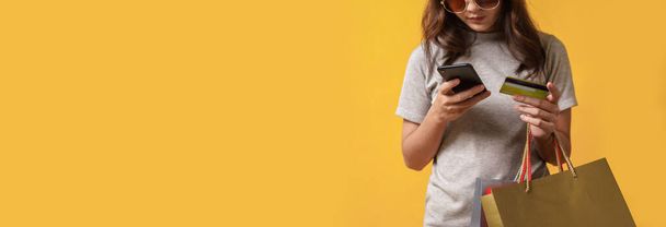 Schöne asiatische Shopaholic Frau trägt Einkaufstüten auf gelbem Hintergrund, während mit dem Smartphone bezahlen online mit einer Kreditkarte. Online-Shopping, Mid-year sales banner concept. Konzentration auf ihre Hand. - Foto, Bild