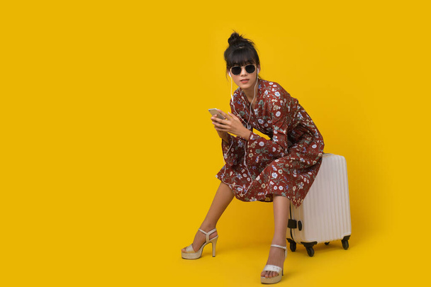 Mladá asijská žena turista poslech hudby ve sluchátku, zatímco sedí na zavazadla na žlutém pozadí studia s kopírovat prostor. - Fotografie, Obrázek