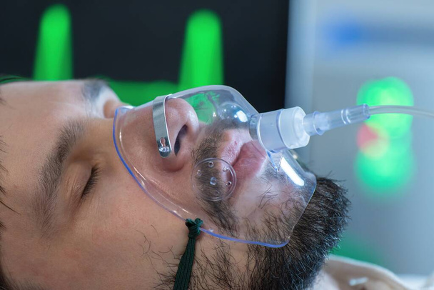 Νεαρός άνδρας αναπνέει με μάσκα οξυγόνου στο πρόσωπο στο νοσοκομείο κατά τη διάρκεια πανδημίας covid-19. - Φωτογραφία, εικόνα