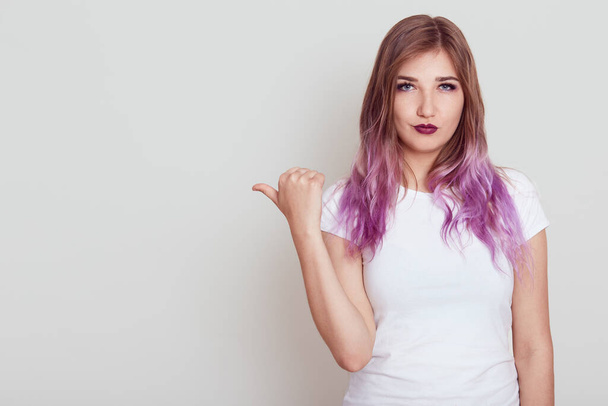 Serious woman wearing casual white t shirt looking at camera with strict expression, pointing thumb aside, showing free space for advertisement, posing isolated over gray background. - 写真・画像
