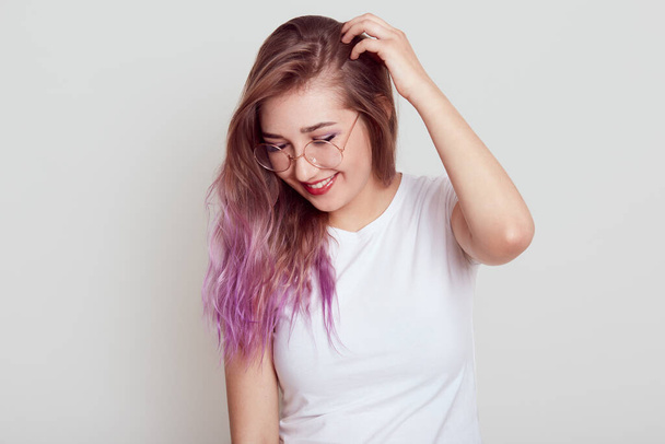 Stylish young girl with lilac hair wearing white t shirt and glasses with positive expression, looking down, keeping hand on her head, dreams, posing isolated over gray background. - Photo, Image