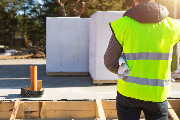 The project architect's back is on the construction site of a house with the foundation laid and the blocks delivered. Construction worker in a protective yellow hardhat and a signal vest. Mock up  - Photo, Image