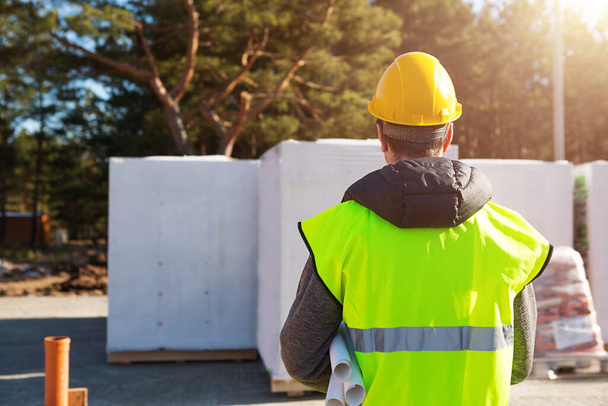 The project architect's back is on the construction site of a house with the foundation laid and the blocks delivered. Construction worker in a protective yellow hardhat and a signal vest. Mock up  - Foto, Bild