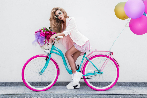 Aantrekkelijke blonde schoonheid op gedecoreerde fiets met bloemen en ballonnen. Voorjaarsconcept. Mooie natuurlijke vrouw in kleurrijke pastelkledij op de fiets. Meisje met fiets op zonnige dag, buiten. Zomertijd. - Foto, afbeelding