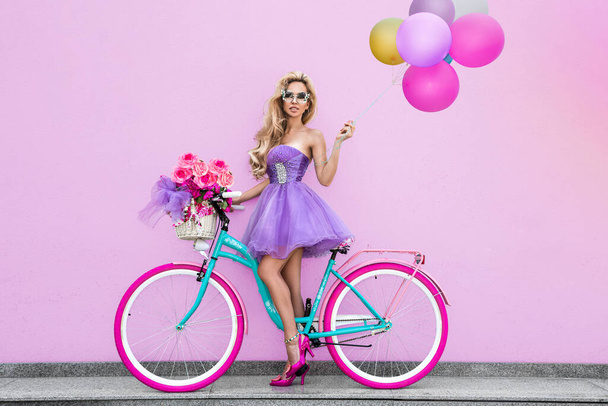 Aantrekkelijke blonde schoonheid op gedecoreerde fiets met bloemen en ballonnen. Voorjaarsconcept. Mooie natuurlijke vrouw in kleurrijke pastelkledij op de fiets. Meisje met fiets op zonnige dag, buiten. Zomertijd. - Foto, afbeelding