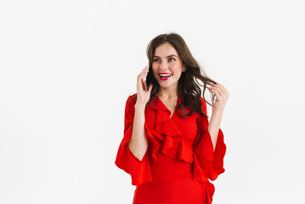 Cheerful young woman in red dress using mobile phone isolated over white background, calling - Zdjęcie, obraz