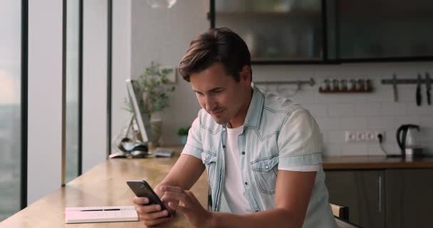 Millennial man sit in domestic kitchen using smartphone - Záběry, video