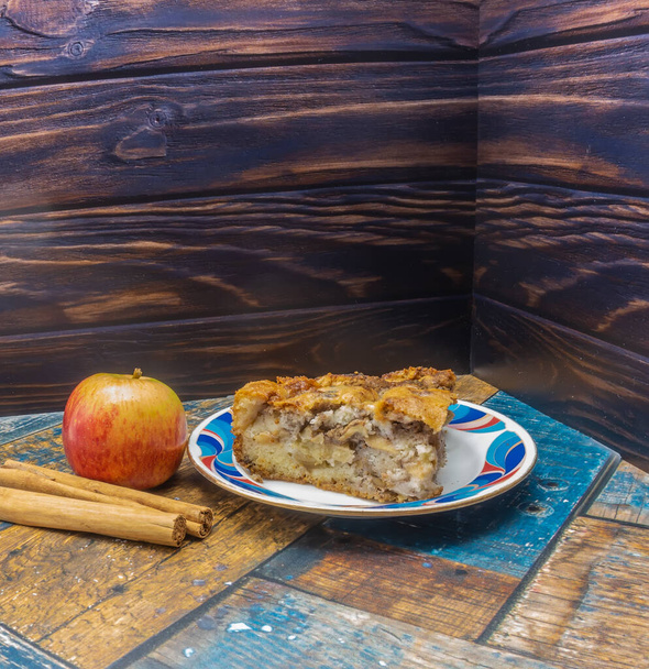 Μια τριγωνική φέτα μηλόπιτας με ξηρούς καρπούς απλώνεται σε ένα πιάτο με ραβδώσεις. Κοντά, στις σανίδες, ένα ώριμο μήλο, ξυλάκια κανέλας. Σπιτικό ψήσιμο. - Φωτογραφία, εικόνα