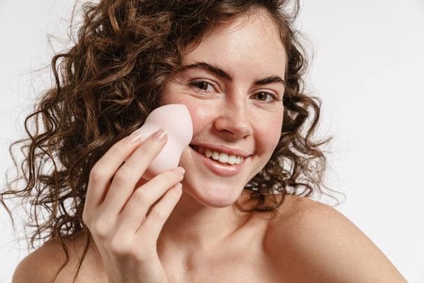 Half-naked curly woman showing cosmetic sponge and smiling at camera isolated over white background - Photo, Image