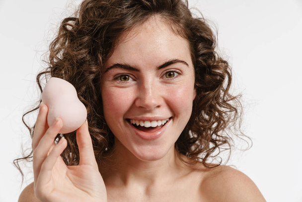 Half-naked curly woman showing cosmetic sponge and smiling at camera isolated over white background - Foto, imagen