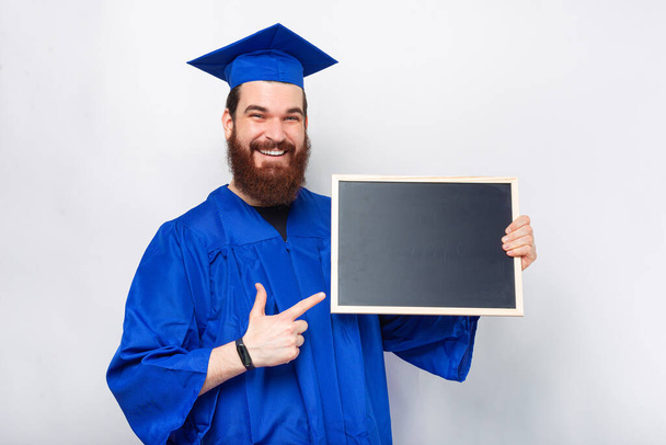 Vrolijke jonge man met baard draagt blauwe vrijgezel en wijst naar leeg zwart krijtbord. - Foto, afbeelding