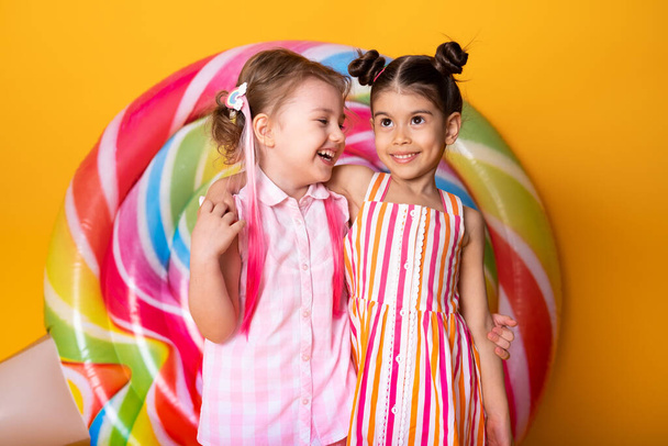 dos niñas felices en vestido colorido riendo abrazos divertirse sobre fondo amarillo con piruleta. - Foto, Imagen