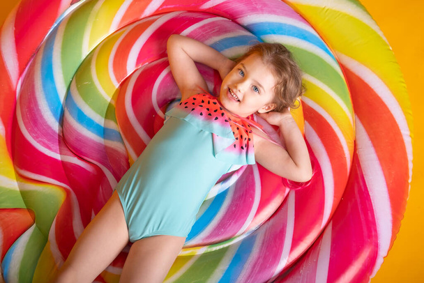 Щаслива маленька дівчинка в купальнику лежить на барвистому надувному матраці, дивлячись на жовтий фон
 - Фото, зображення