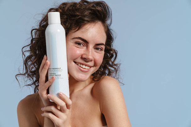 Shirtless curly woman smiling and showing facial mist isolated over blue background - Foto, Bild