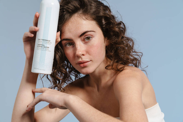 European curly woman posing and showing facial mist isolated over blue background - Foto, Imagem