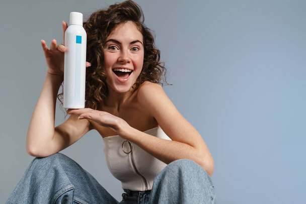 Ενθουσιασμένη σγουρή κοπέλα που δείχνει ιαματικό νερό ενώ κάθεται απομονωμένη σε μπλε φόντο - Φωτογραφία, εικόνα