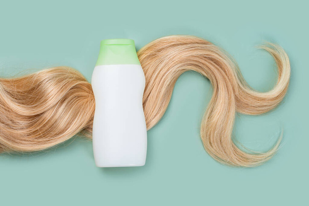Shampoo or conditioner bottle wrapped in lock of curly blonde hair on light mint background, top view. Flat lay in pastel colors, mockup. Hair care cosmetics, beauty haircare products, hair treatment - Foto, afbeelding