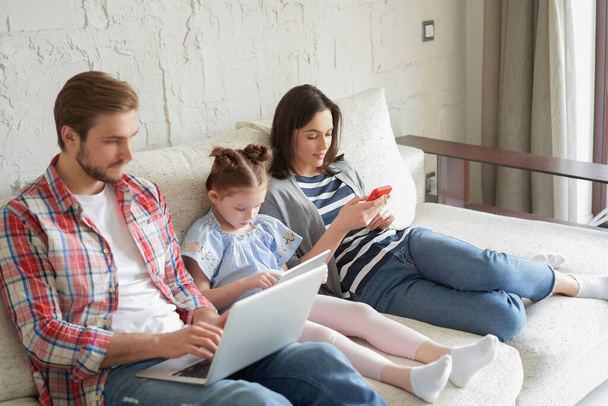 Father, mother and daughter using electronic devices sitting on sofa at living room - Фото, изображение