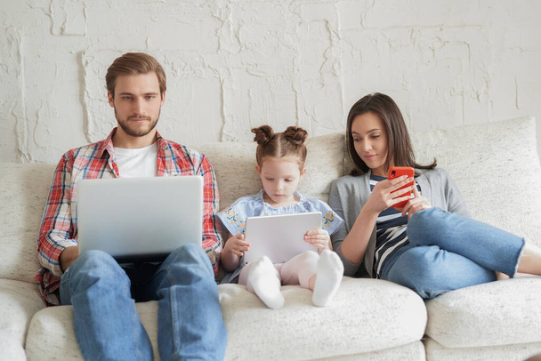 Padre, madre e hija usando dispositivos electrónicos sentados en el sofá en la sala de estar - Foto, Imagen