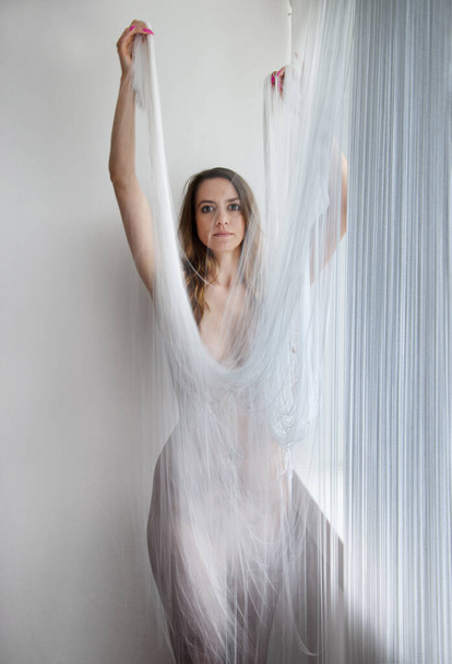 μυώδης όμορφη νεαρή ελκυστική σέξι γυναίκα από το παράθυρο, σε ένα σώμα κολακευτικό μοντέρνο κινούμενο φόρεμα από τα λευκά νήματα μιας πτυσσόμενης κουρτίνας κλωστής - Φωτογραφία, εικόνα
