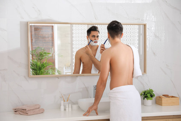 Handsome young man shaving with razor near mirror in bathroom - Fotoğraf, Görsel