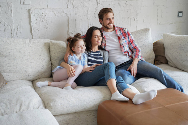 Beautiful happy young parents and their little daughter watching TV and smiling while sitting on sofa at home - Valokuva, kuva