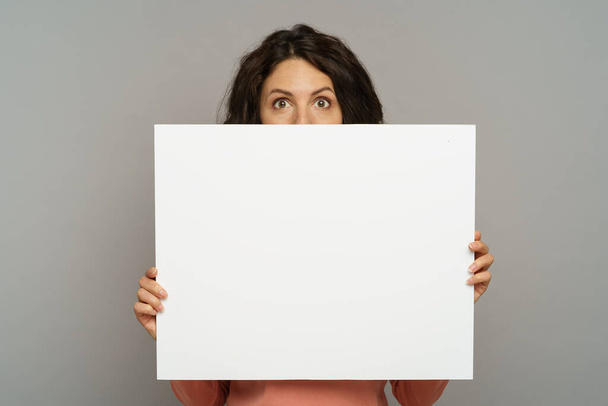 Junge brünette Frau halten Plakatwand isoliert auf grauem Hintergrund. Millennial Mädchen zeigen leeres Plakat - Foto, Bild