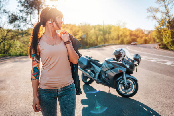 道路でバイクの背景に革のジャケットを着たかなり若い入れ墨の女性がポーズをとります.オートバイ旅行のコンセプト. - 写真・画像