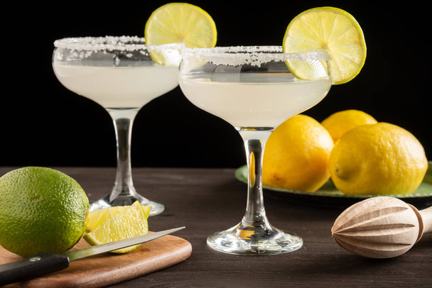 Close-up of two glasses with margarita cocktail, on dark wooden table with limes and lemons, black background, horizontal - Photo, image