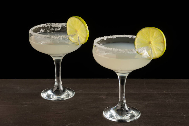 Top view of two glasses of margarita cocktail with lime slices and salt, on wooden table, horizontal black background, with copy space - Photo, image