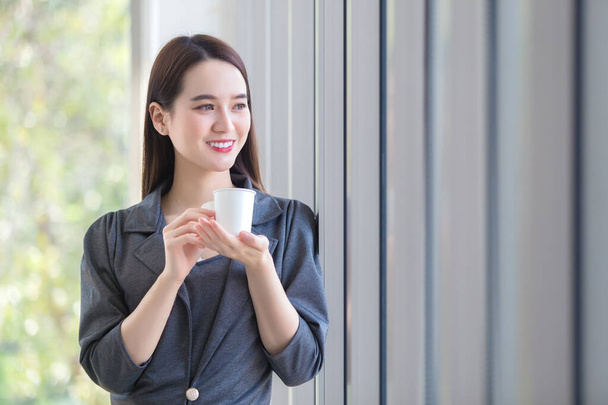 Aasialainen työssäkäyvä nainen katsoo ulos ikkunasta ajatella jotain samalla kun hän pitelee paperista tehtyä kahvikuppia toimistossa. - Valokuva, kuva