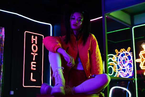 brunette young asian woman sitting near neon sign with hotel lettering outside  - Foto, imagen
