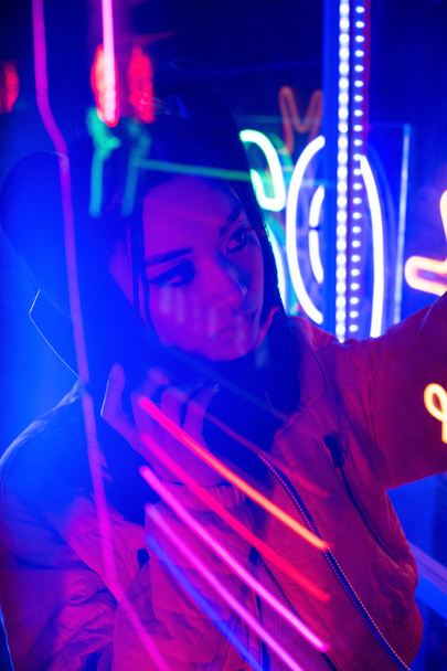 neon lighting near young asian woman talking on retro payphone outside  - 写真・画像