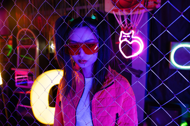 trendy young asian woman in red sunglasses near neon sign and metallic fence  - Fotografie, Obrázek