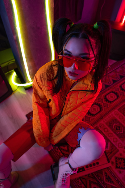high angle view of stylish young asian woman in sunglasses and orange jacket posing near neon lighting  - Zdjęcie, obraz