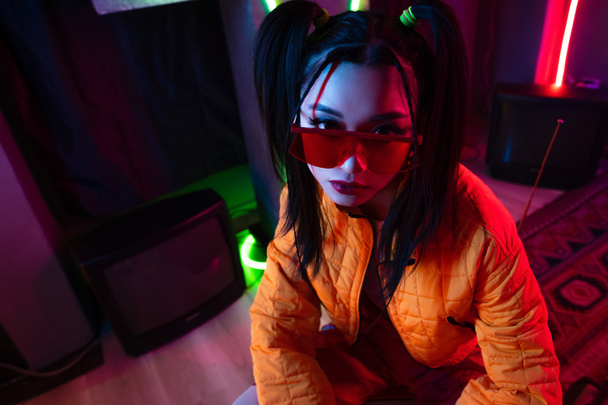 high angle view of stylish young asian woman in sunglasses and orange jacket posing near neon lighting  - 写真・画像
