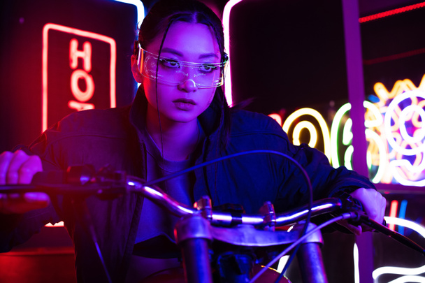 молодая азиатка в солнечных очках едет на мотоцикле с неоновым освещением позади  - Фото, изображение